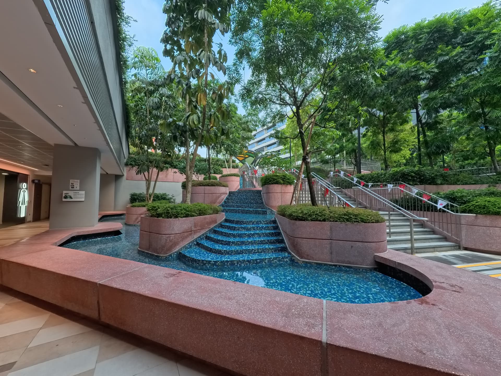 Stepped water cascade at the Amphitheatre on level B1, flanked by planters and open to the sky, with the covered corridor alongside