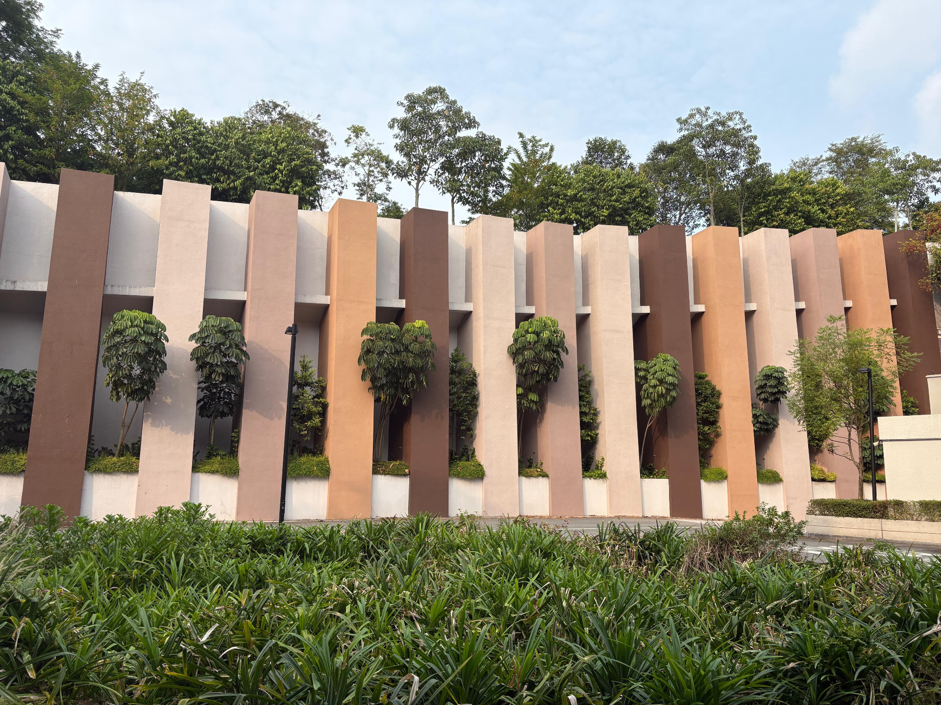 A boundary wall of staggered vertical fins in terracotta and cream, with trees planted in the ground between the fins and a continuous planter at the base