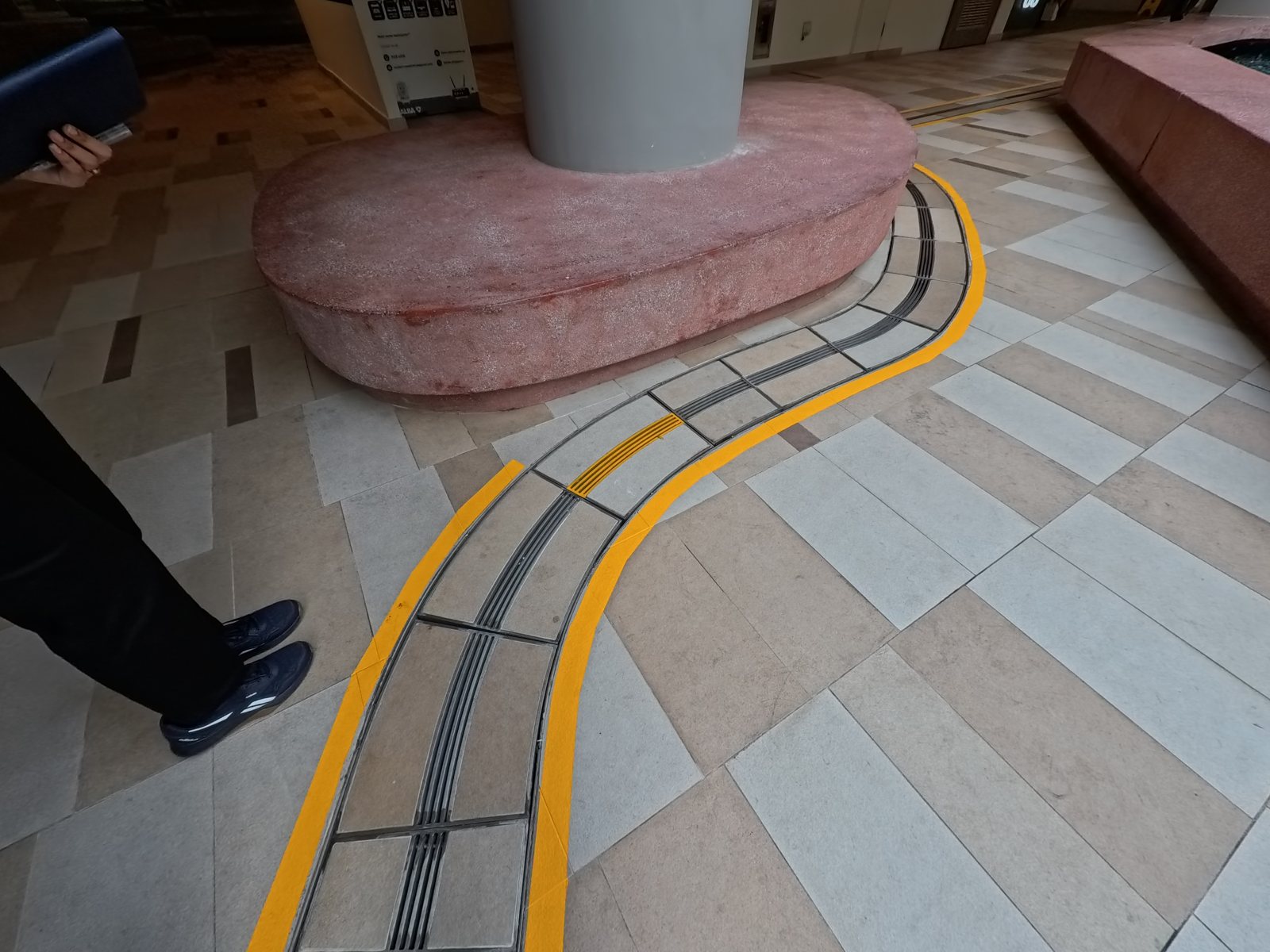 A linear channel drain with dark grating set into a tiled floor under cover, curving around a pink terrazzo bench and a column, flanked on both sides by painted yellow lines.