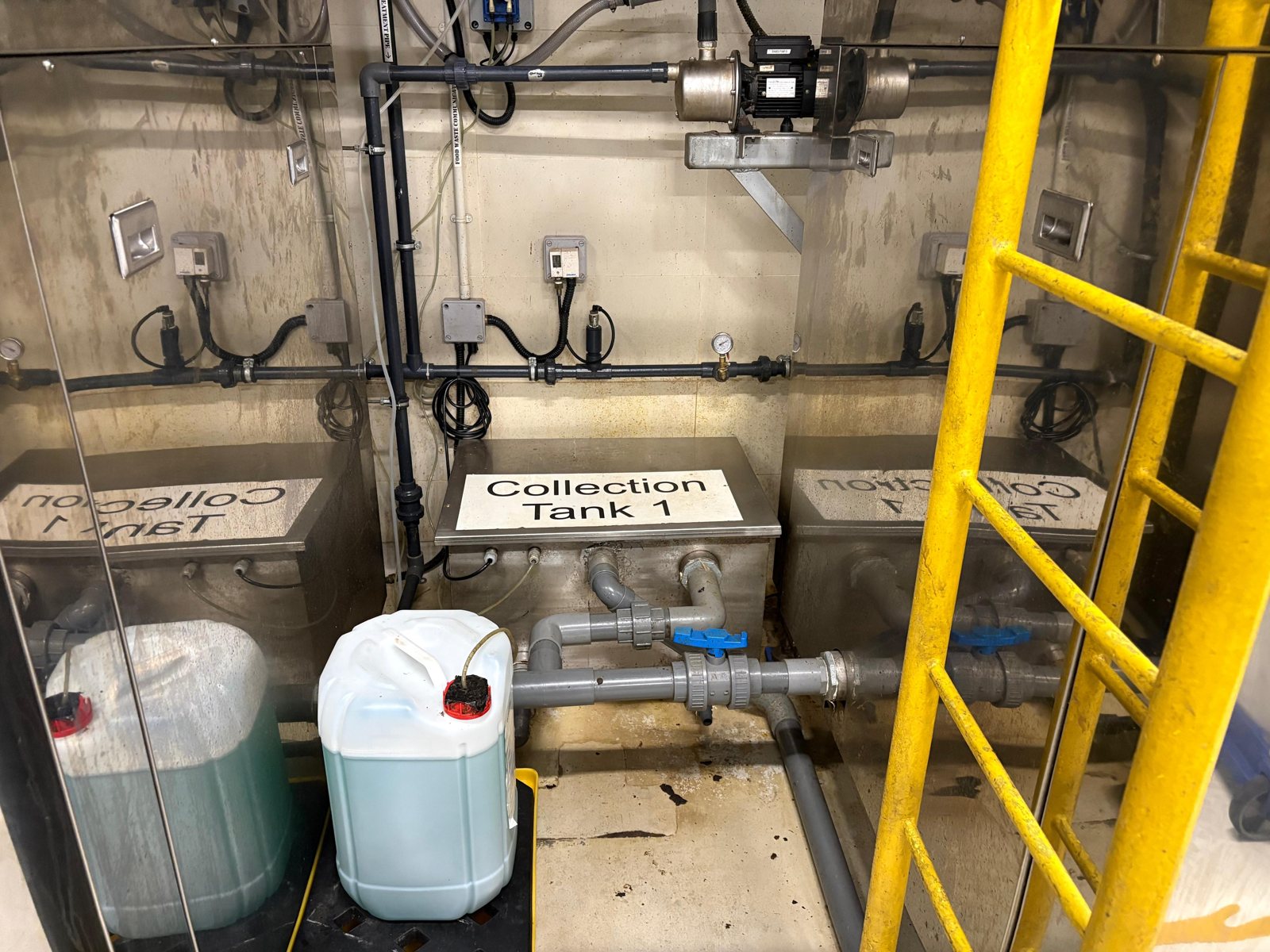 A plant room with a stainless steel vessel labelled Collection Tank 1, pipework, pressure gauges, a wall mounted pump, and a plastic drum on the floor with a dosing tube through its cap. Wall signage reads Food Waste Comminutor and Treatment Pipe.